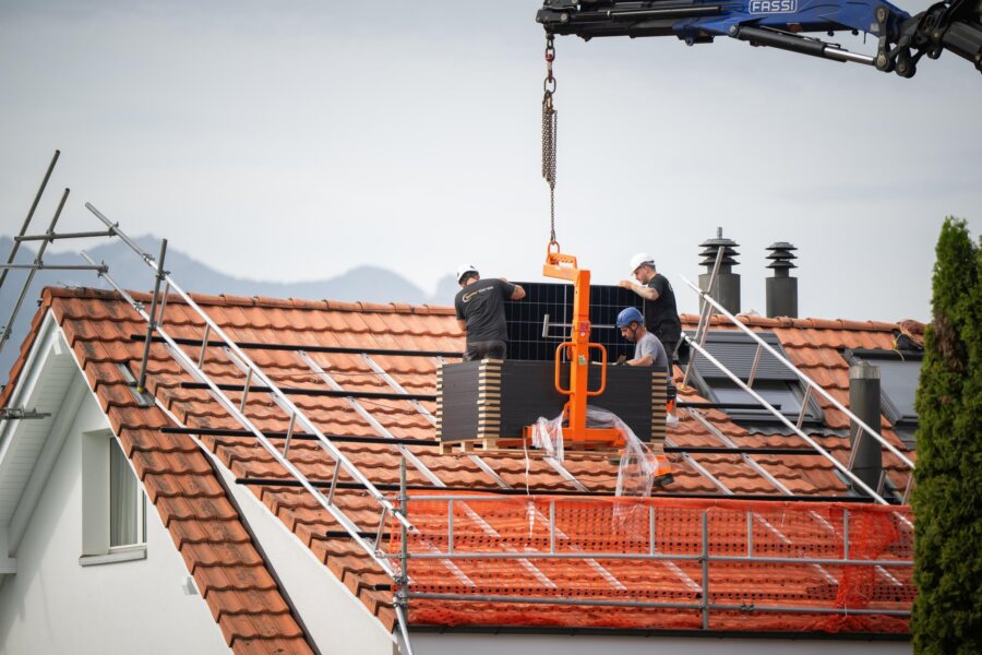 Ein Kran setzt ein Paket Photovoltaik-Module auf dem Ziegeldach ab, zwei Monteure warten darauf.