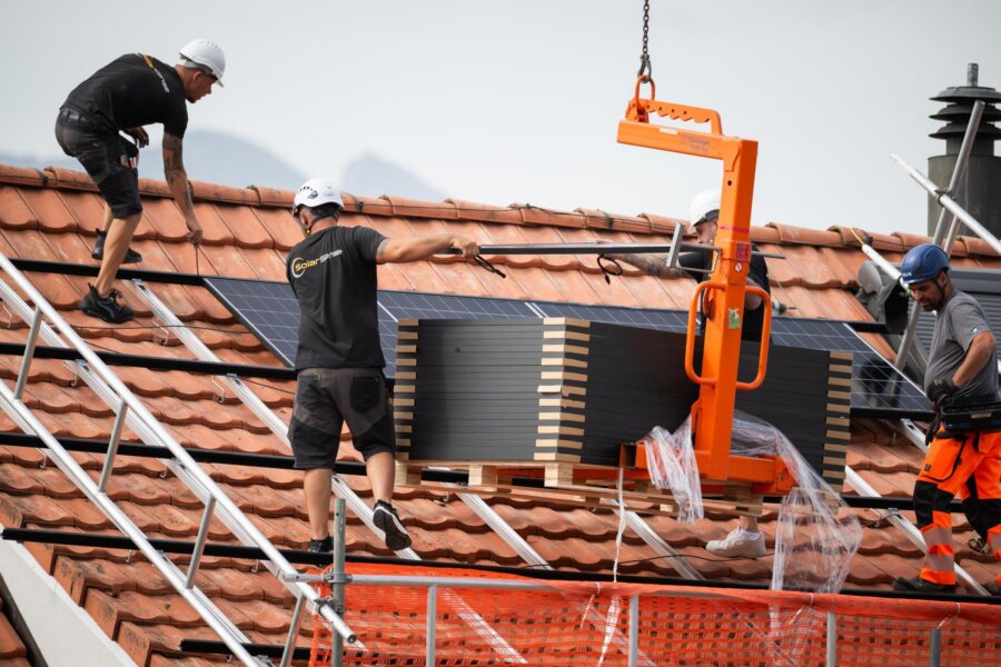 Zwei Monteure verlegen Module auf einem Ziegeldach, daneben der orange Modulheber.