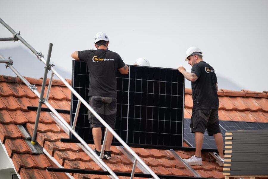 Zwei Monteure tragen ein Photovoltaik-Modul über das Gerüst auf dem Ziegeldach.