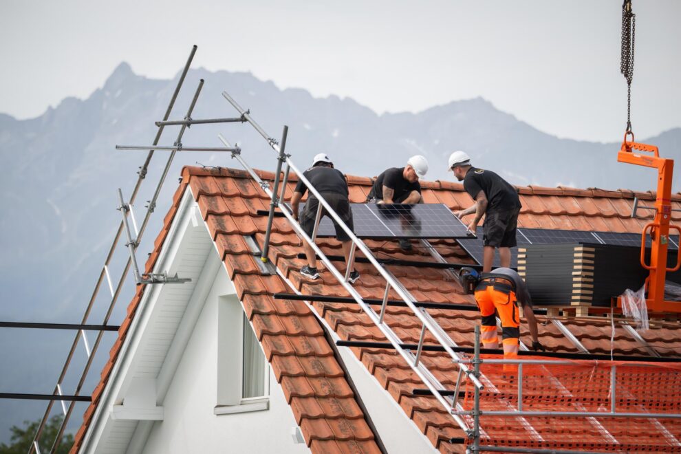 Drei Monteure arbeiten auf einem Ziegeldach an der neuen Photovoltaik-Anlage, im Hintergrund die Berge.