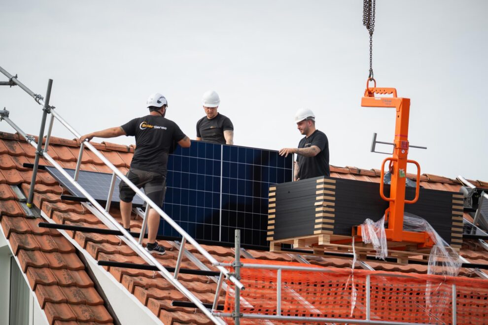 Drei Monteure stellen auf dem Dach ein Photovoltaik-Modul auf, der Kranhaken hängt daneben.