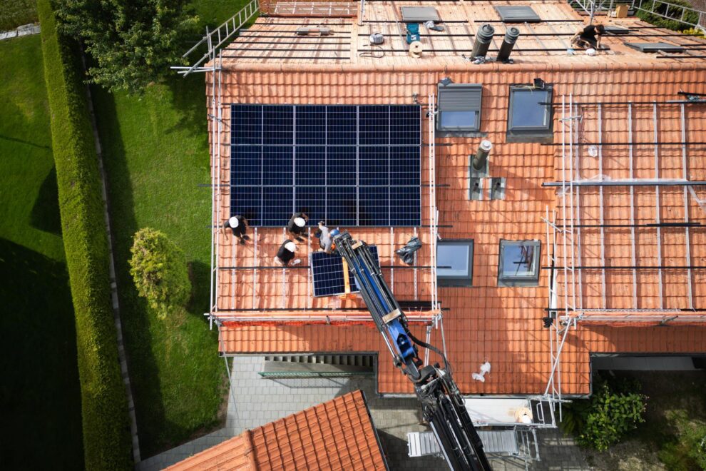 Luftaufnahme des ganzen Hauses mit der halb fertigen Photovoltaik-Anlage und dem Kranwagen davor.